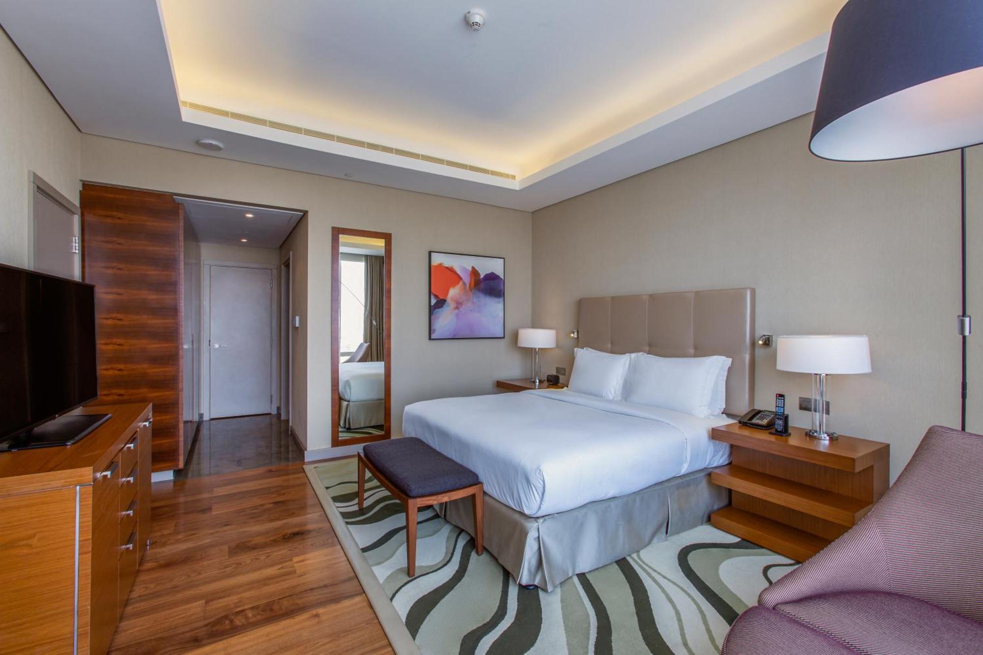 Hilton Riyadh Hotel & Residences エクステリア 写真 The photo shows a modern hotel room with a comfortable and stylish design. It features a large bed with white linens and a headboard, flanked by two bedside lamps. There is a small bench at the foot of the bed and a cozy chair in the corner. A flat-s