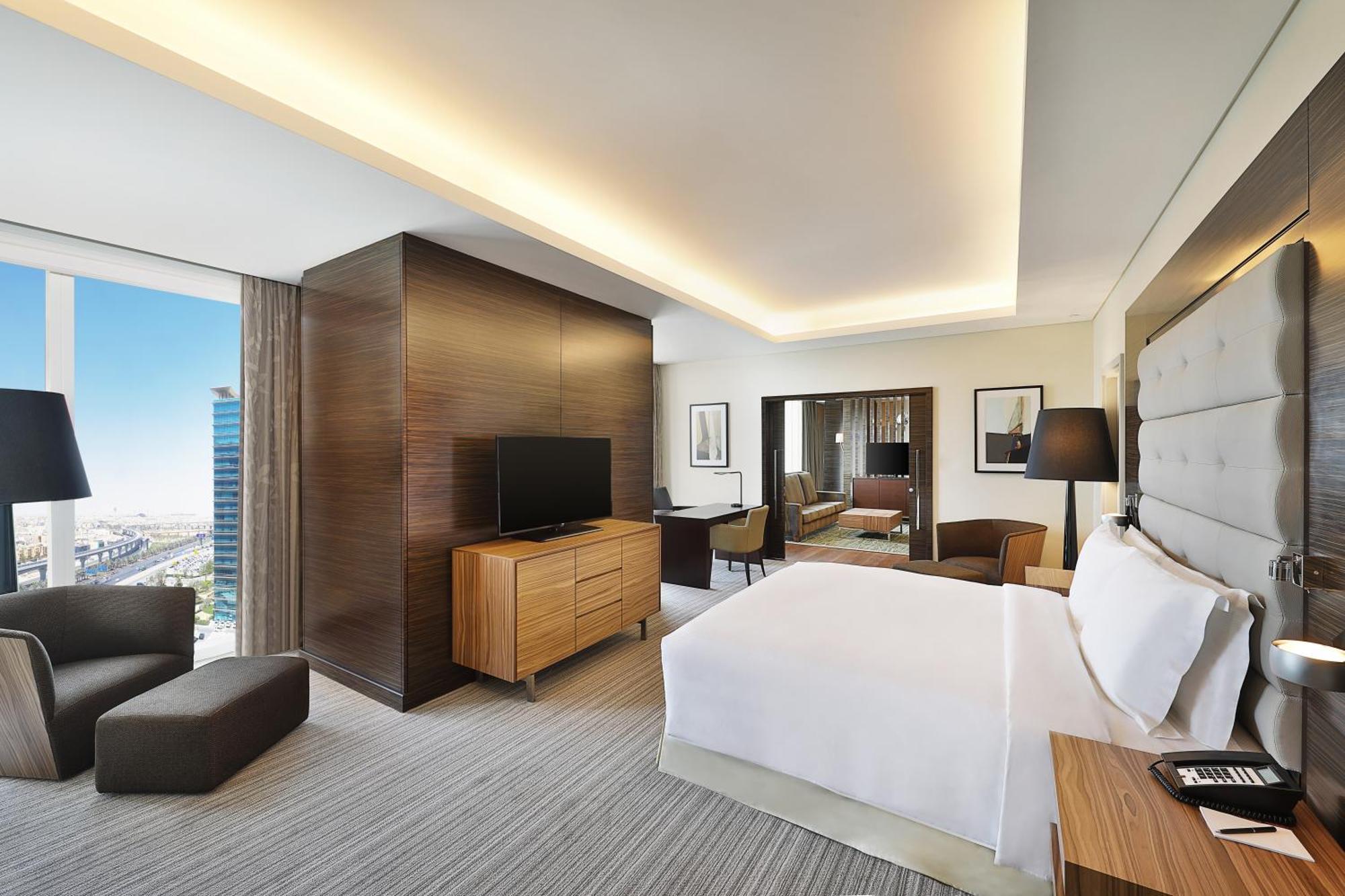 Hilton Riyadh Hotel & Residences エクステリア 写真 The photo shows a modern hotel room. It features a neatly made bed with white linens, a wooden nightstand beside it, and a cozy armchair. The room has a large window that provides a view of the outside, likely showcasing a cityscape. A flat-screen TV
