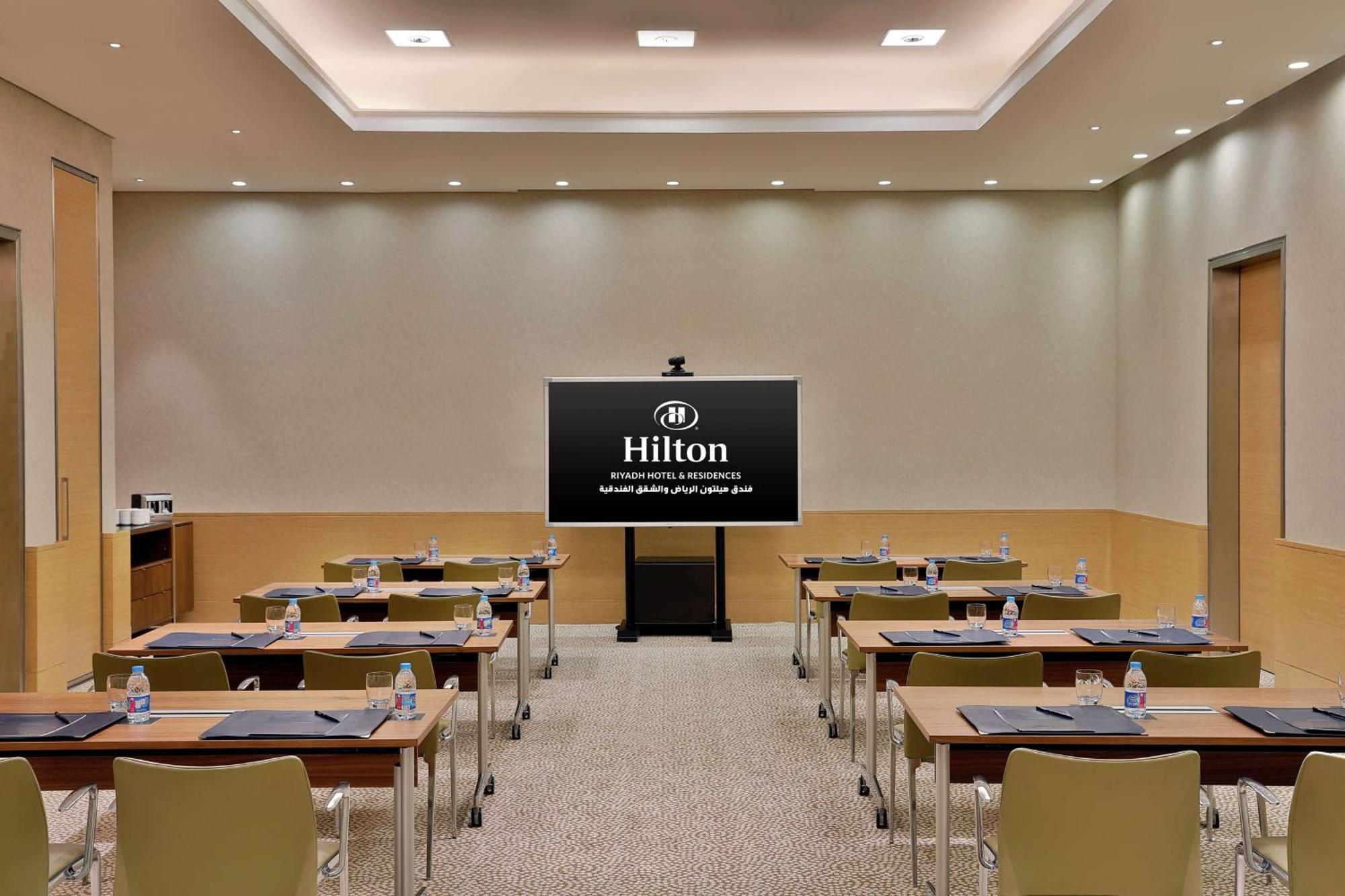 Hilton Riyadh Hotel & Residences エクステリア 写真 The photo shows a conference or meeting room set up for a presentation. There are several tables arranged in a classroom style, each equipped with chairs. On each table, there are notepads and water bottles. At the front of the room is a large screen
