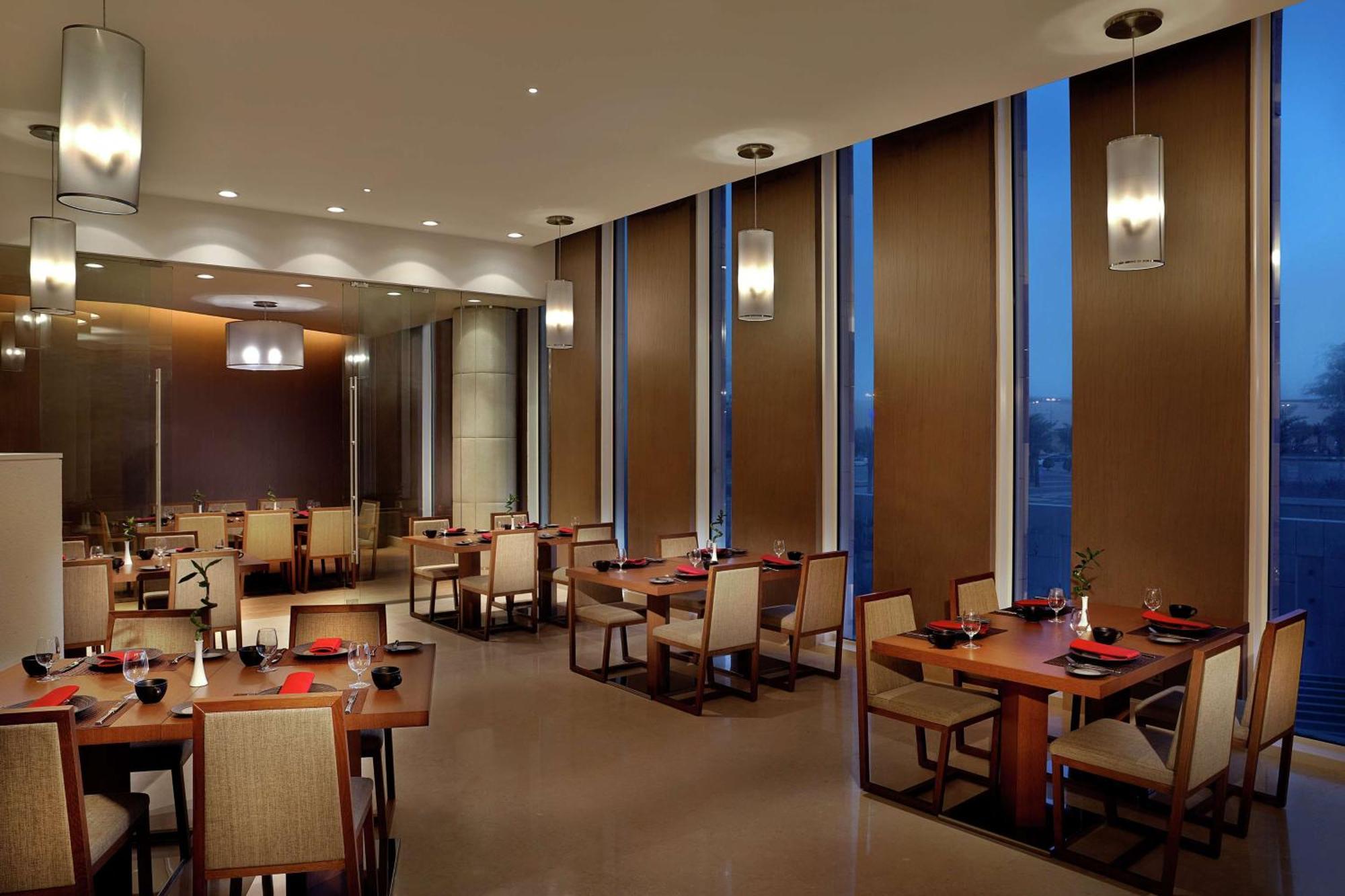 Hilton Riyadh Hotel & Residences エクステリア 写真 The image depicts a modern restaurant interior. It features several dining tables arranged neatly, each set with plates and utensils. The room has large windows with blinds partially drawn, allowing some ambient light to filter in, while evening ligh
